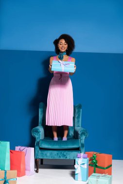 smiling african american woman standing on velour armchair with gift box near purchases on blue background