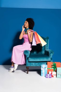 smiling african american woman sitting in velour armchair with purchases and talking on smartphone near gifts on blue background
