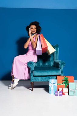 smiling african american woman sitting in velour armchair with purchases and talking on smartphone near gifts on blue background