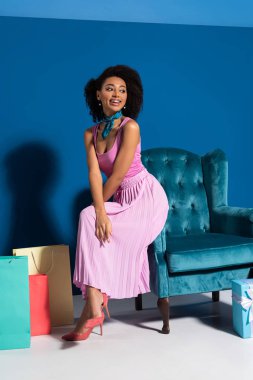 smiling african american woman sitting on velour armchair near gift boxes and purchases on blue background