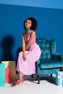 african american woman sitting on velour armchair near gift boxes and purchases on blue background