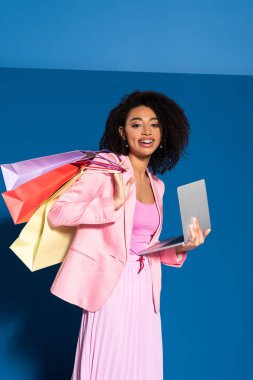 smiling elegant african american businesswoman with shopping bags using laptop on blue background