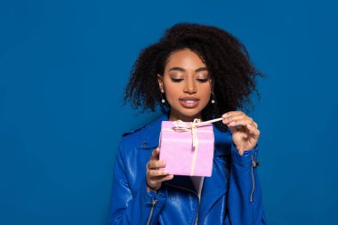 smiling african american woman opening gift isolated on blue background