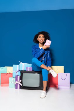 smiling african american woman sitting on vintage television near gifts and shopping bags on blue background