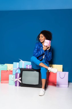 smiling african american woman sitting on vintage television near gifts and shopping bags on blue background