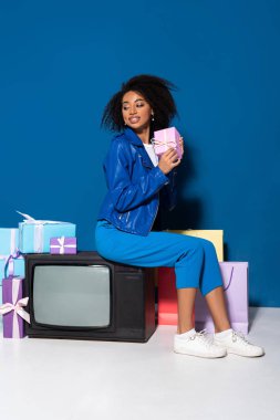 smiling african american woman sitting on vintage television near gifts and shopping bags on blue background