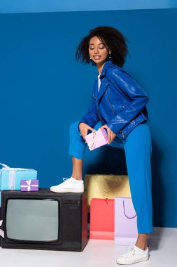smiling african american woman standing on vintage television near gifts and shopping bags on blue background