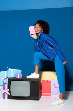 smiling african american woman standing on vintage television with gift on blue background