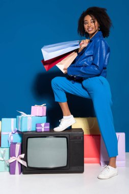 smiling african american woman sitting on vintage television with shopping bags on blue background