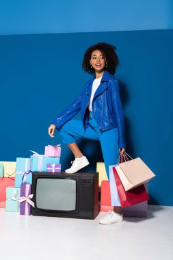 smiling african american woman sitting on vintage television with shopping bags on blue background