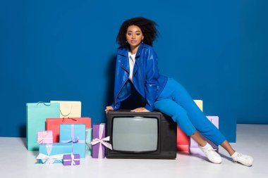 african american woman sitting on vintage television near gifts and shopping bags on blue background