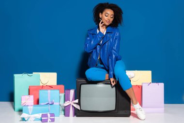 smiling african american woman sitting on vintage television near gifts and shopping bags on blue background