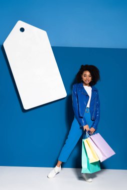 smiling african american woman with shopping bags near big blank price tag on blue background