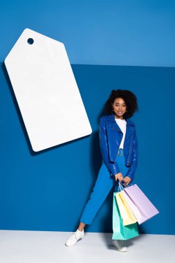 smiling african american woman with shopping bags near big blank price tag on blue background
