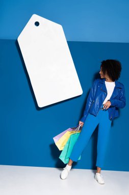 smiling african american woman with shopping bags near big blank price tag on blue background