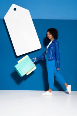 african american woman walking with shopping bags near big blank price tag on blue background