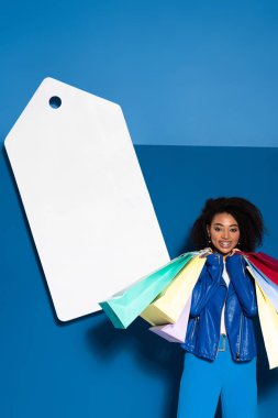smiling african american woman with shopping bags near big blank price tag on blue background