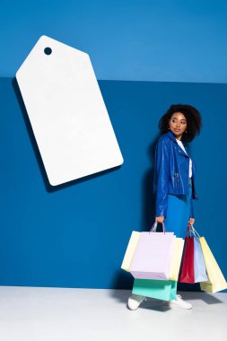 smiling african american woman with shopping bags near big blank price tag on blue background