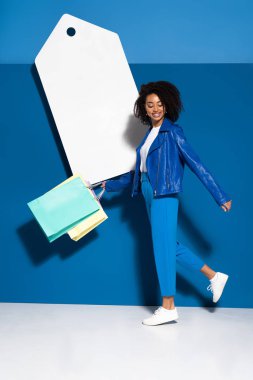 happy african american woman walking with shopping bags near big blank price tag on blue background