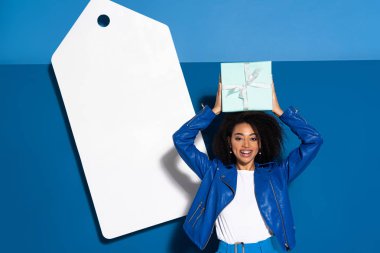 smiling african american woman with gift near big blank price tag on blue background