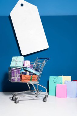 shopping bags, trolley with gifts and big blank price tag on blue background
