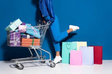 cropped view of african american woman with shopping cart full of gifts near shopping bags on blue background