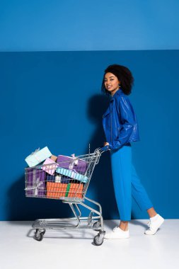 side view of happy african american woman with shopping cart full of gifts on blue background