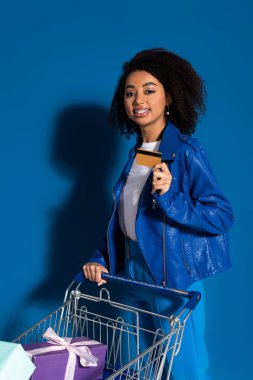 happy african american woman with credit card and shopping cart full of gifts on blue background