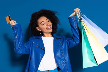 smiling african american woman with shopping bags and credit card on blue background