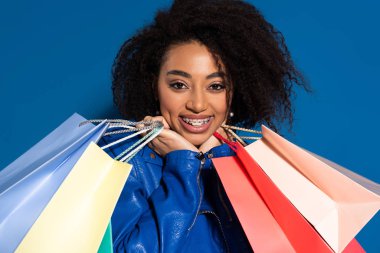smiling african american woman with braces and shopping bags isolated on blue