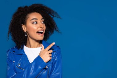 happy african american woman with open mouth pointing with finger aside isolated on blue