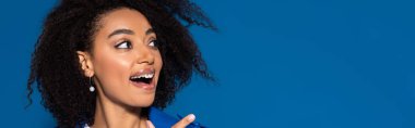 happy african american woman with open mouth pointing with finger aside isolated on blue, panoramic shot