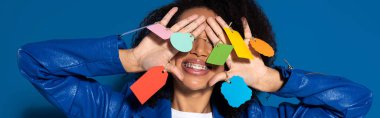 smiling african american woman covering eyes with empty labels on hands on blue background, panoramic shot