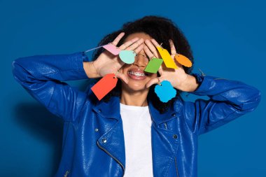 smiling african american woman covering eyes with empty labels on hands on blue background