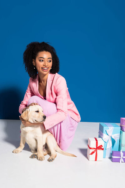 улыбающаяся африканская американка с золотистым щенком ретривера рядом с подарками на синем фоне
