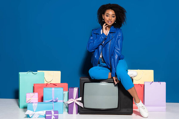 улыбающаяся африканская американка, сидящая на винтажном телевизоре рядом с подарками и пакетами для покупок на синем фоне
