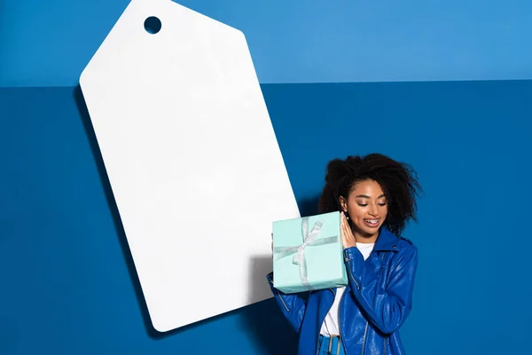 Улыбающаяся Африканская Американка Подарком Рядом Большим Пустым Ценником Синем Фоне — стоковое фото
