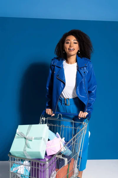 smiling african american woman with shopping cart full of gifts on blue background