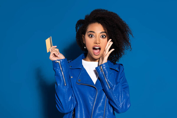 shocked african american woman with credit card on blue background