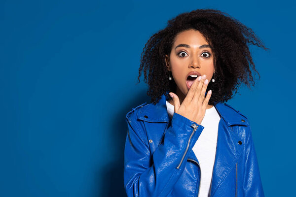 shocked african american woman coveting mouth with hand on blue background