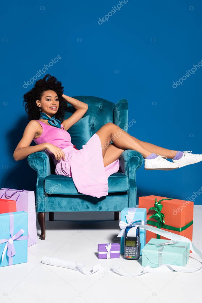 smiling african american woman sitting in velour armchair near payment terminal with check, shopping bags and gifts on blue background