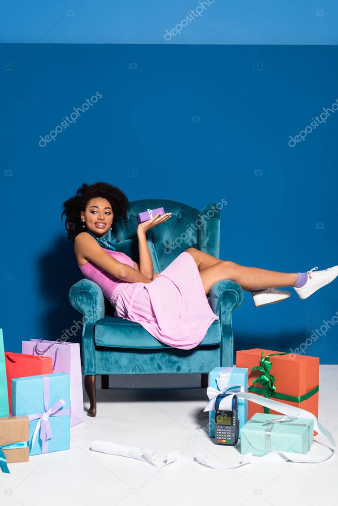 smiling african american woman sitting in velour armchair with gift box near purchases on blue background