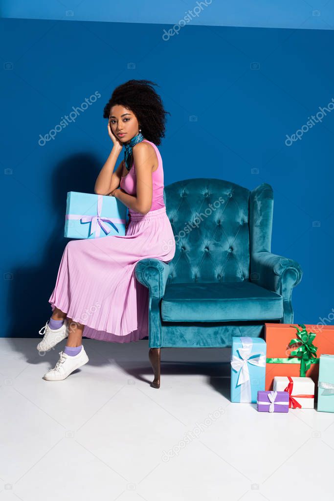 african american woman sitting on velour armchair near gift boxes on blue background