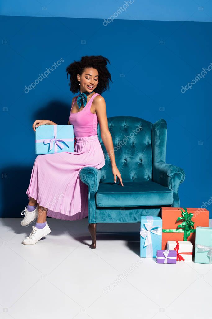 smiling african american woman sitting on velour armchair near gift boxes on blue background
