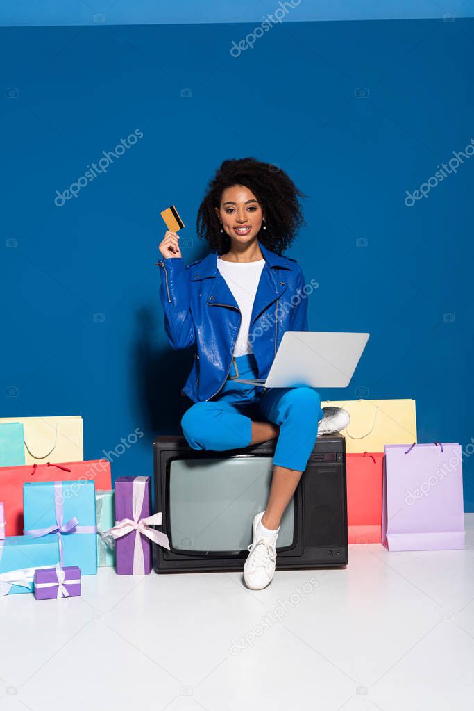 smiling african american woman sitting on vintage television with laptop and credit card near gifts and shopping bags on blue background