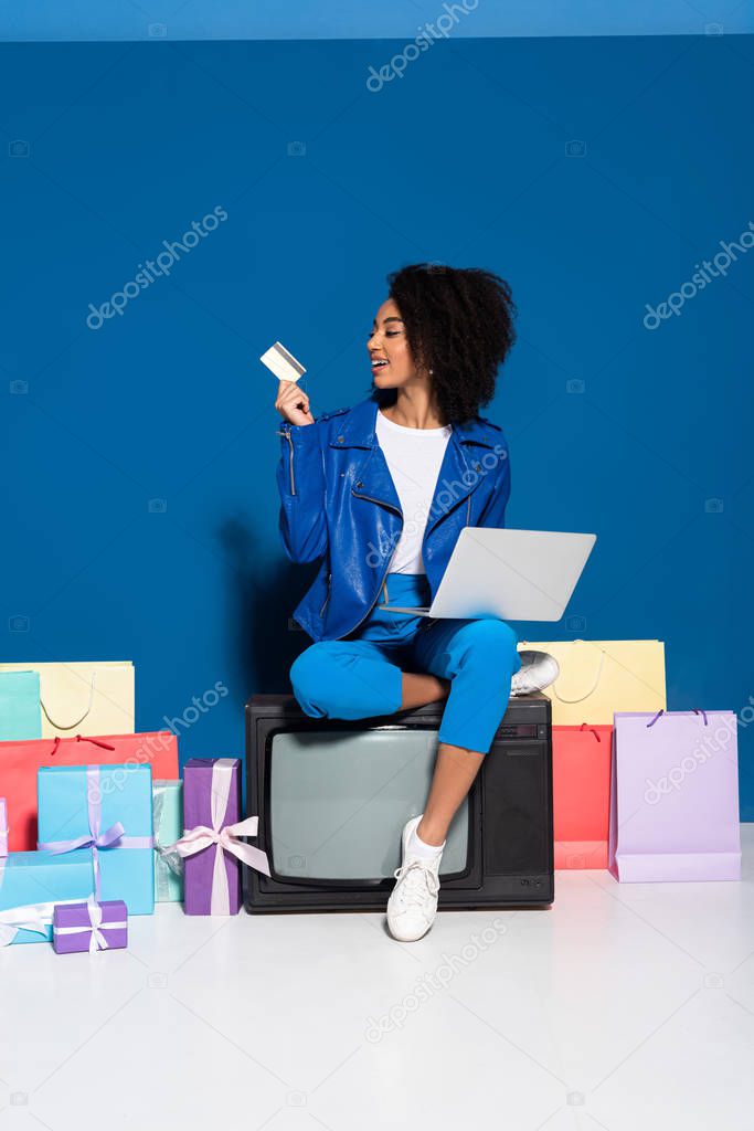 smiling african american woman sitting on vintage television with laptop and credit card near gifts and shopping bags on blue background