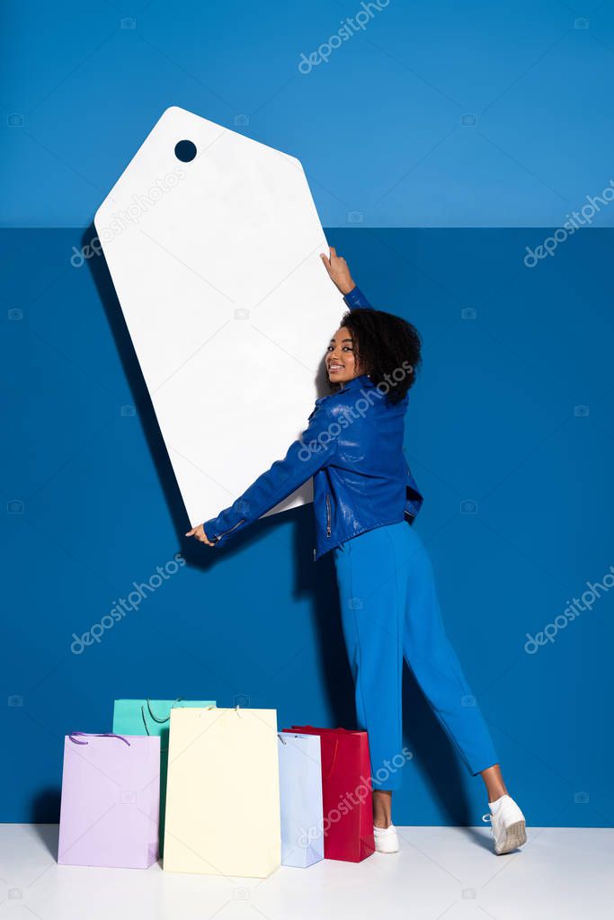 smiling african american woman holding big blank price tag near shopping bag on blue background