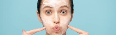 panoramic shot of funny woman with cleansing foam on face pointing on cheeks, isolated on blue clipart