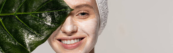 panoramic shot of attractive smiling girl with cosmetic mask on face posing with green leaf, isolated on grey 