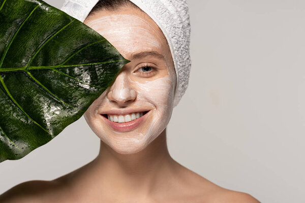 happy young woman with cosmetic mask on face and towel on head posing with green leaf, isolated on grey 
